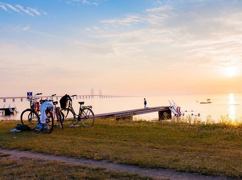 Cyklar i kvällsljuset på Sibbarpsstranden med en badbrygga och Öresundsbron i bakgrunden