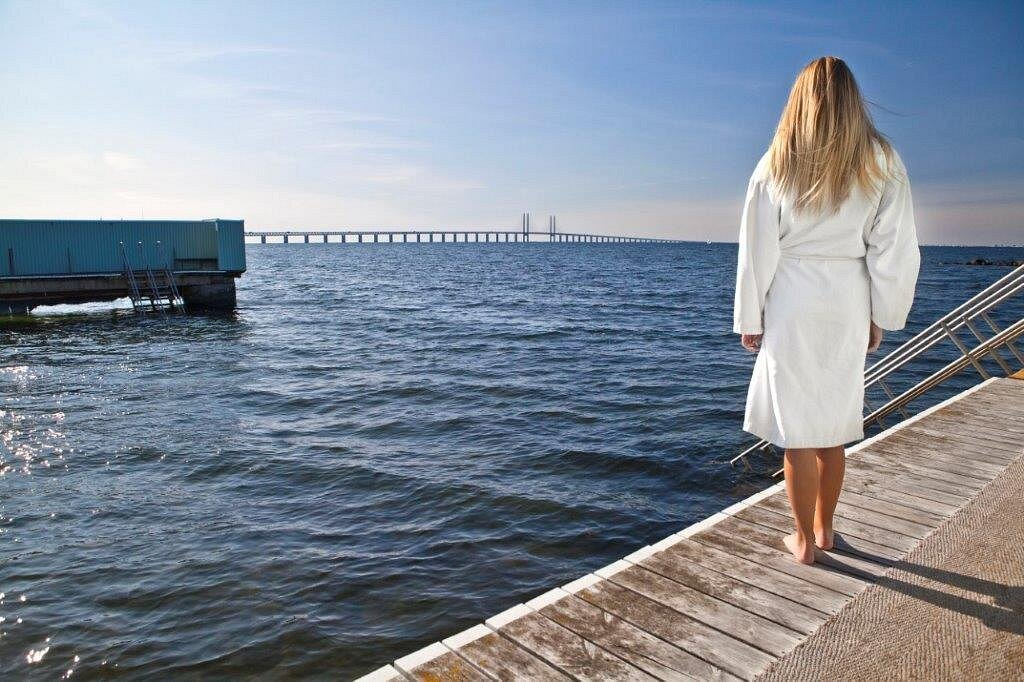 Besökare i vit badrock vid Sibbarps kallbadhus med Öresundsbron i bakgrunden
